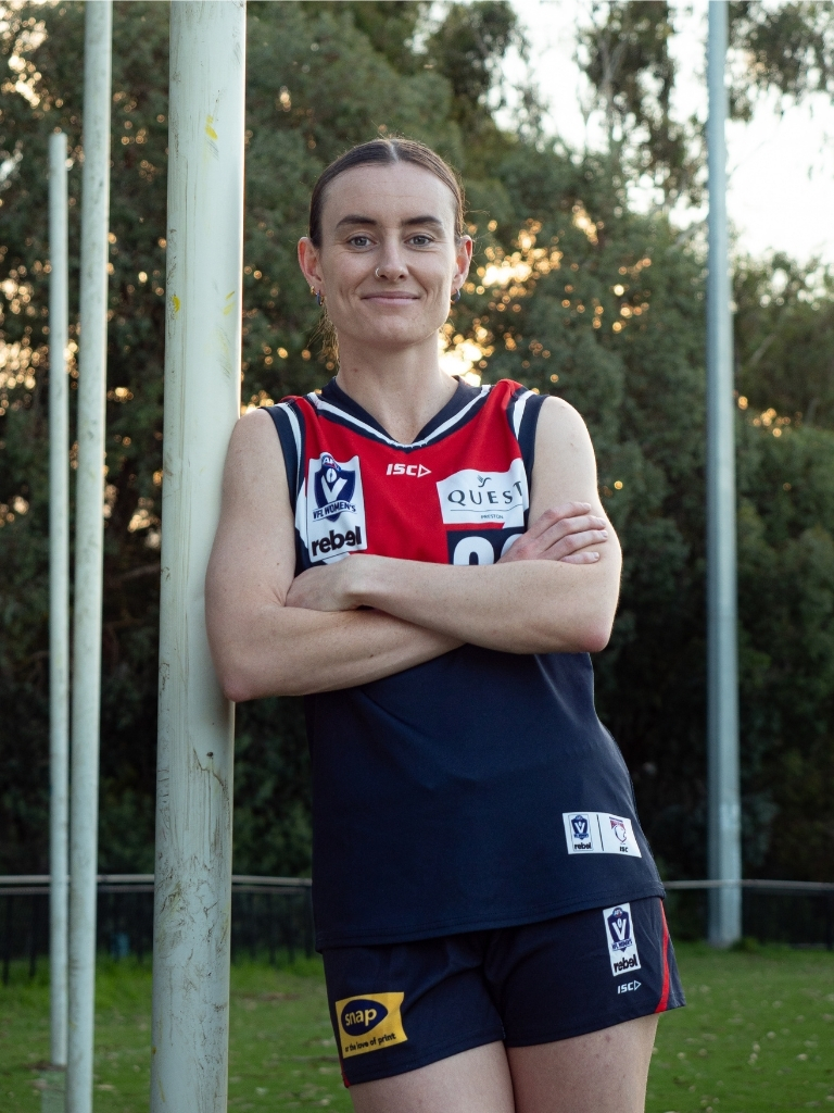 Caitlin Bunker stands leaning against a goal post with arms crossed, wearing the Darebin Falcons VFLW guernsey, with the oval and trees visible in the background.